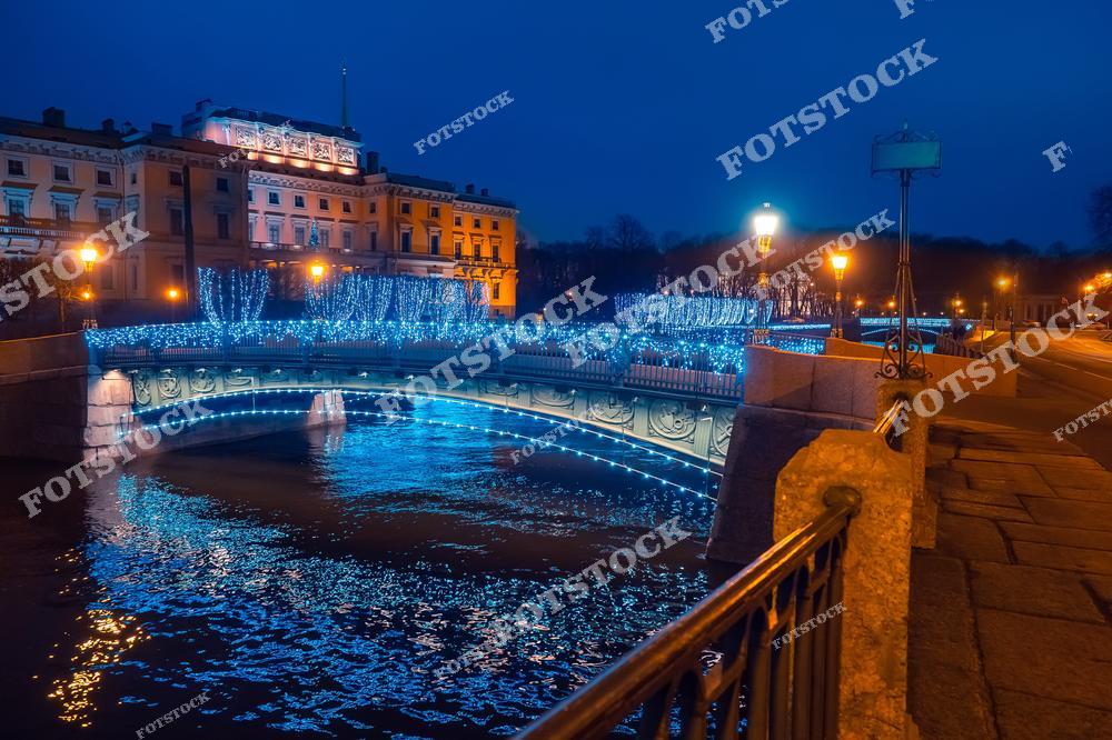 На фотографии изображён Инженерный замок на фоне ярко украшенного моста в Санкт-Петербурге. Мост переливается голубыми огнями, подчёркивая праздничную атмосферу города. Вечернее освещение и отражения в канале создают атмосферу уюта и волшебства. Архитектура и декорации идеально сочетаются, подчеркивая богатую историю города.
